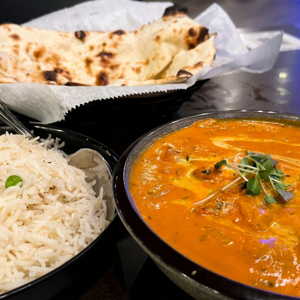 Indian meal featuring basmati rice, creamy orange curry garnished with microgreens, and naan bread served on a dark table.